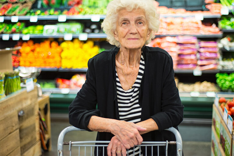 An older woman leans her arms on a shopping cart in the grocery store