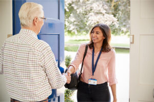 An elderly man stands at the front door of his home, greeting his new caregiver with a handshake and a smile.