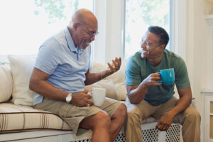 Two smiling men sit together holding mugs, as the younger man uses tips to break conversation loops in dementia with the older man.