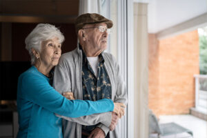 An older woman hugs her husband as they both look out the window, as she knows what to do when you resent caregiving for your spouse.