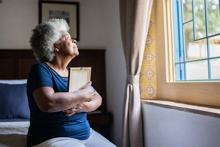 An older woman experiencing feelings of grief and loss clutches a photo to her chest while gazing sadly out the window.