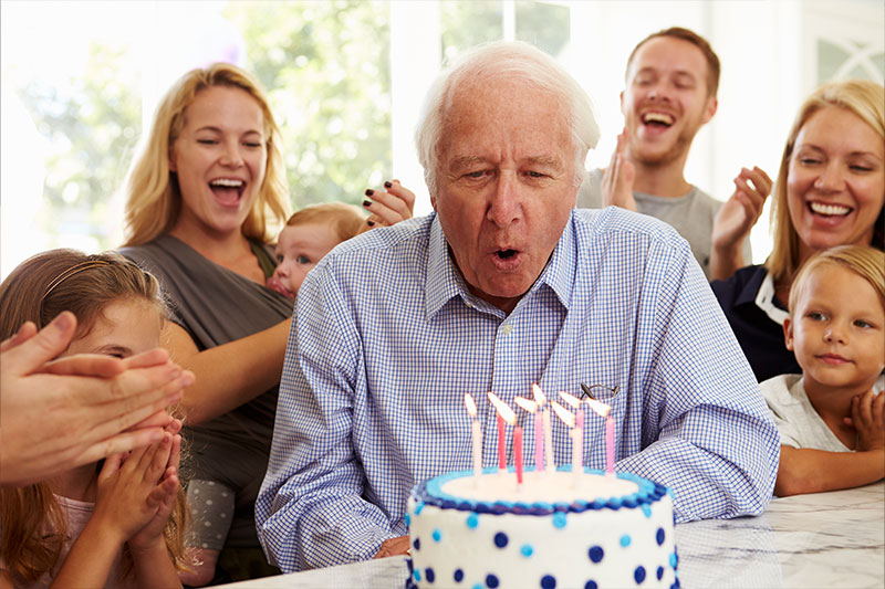 A family who has been planning a senior birthday party smiles and cheers as their older loved one blows out the candles on his cake.