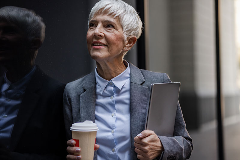 A woman who provides care for aging parents holds a cup of coffee and smiles at work.