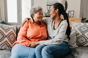 A woman who understands the importance of promoting independence in supporting your aging parents wraps her arm around her mom as they sit on the sofa and smile at each other.