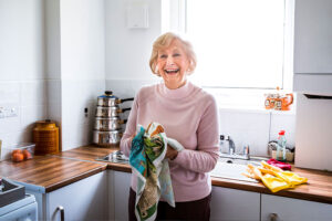 A woman smiles confidently while drying a dish in the kitchen, thanks to her caregiver who understands the need for self-sufficiency for someone with dementia.