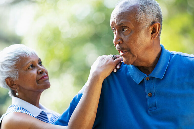 A woman puts her hands on the shoulder of her husband who sometimes struggles with dementia impulsivity.