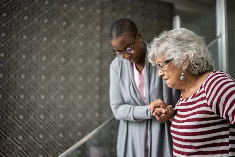 A caregiver who has recognized early signs of dementia in a senior client helps her walk down the stairs.