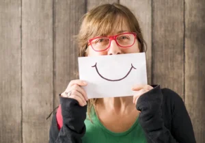 A woman holds a smiley face drawn on a piece of paper over her own mouth to indicate that she’s happy despite the fact that she’s experiencing dementia caregiver stress.